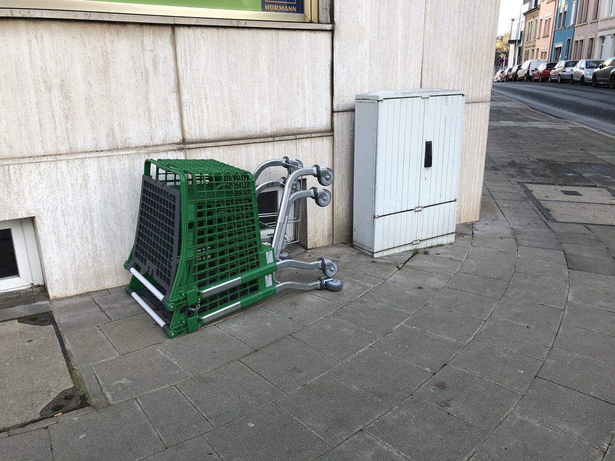 Deux caddies en plastique vert emboîtés l’un dans l’autre sont posés sur le sol sur un trottoir, à côté d’un relais électrique